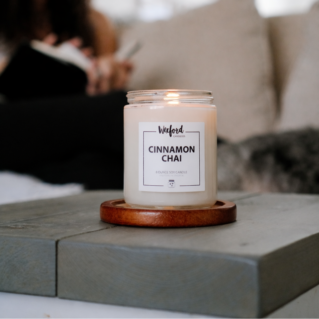 Candle labeled 'Cinnamon Chai' on a wooden coaster with a blurred person and dog in the background.
