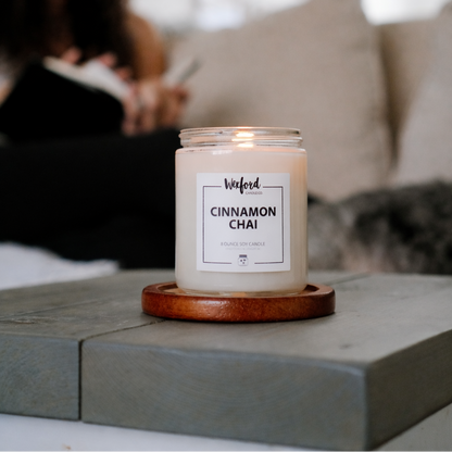 Candle labeled 'Cinnamon Chai' on a wooden coaster with a blurred person and dog in the background.