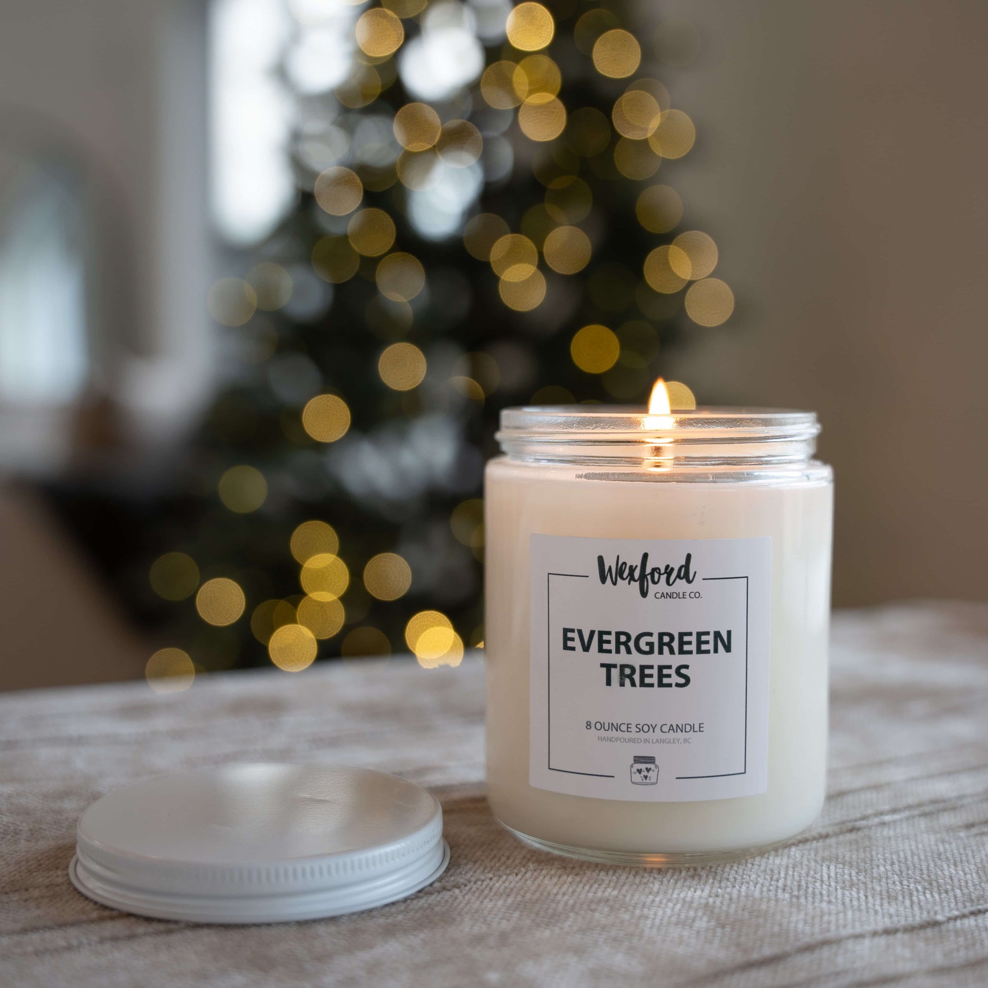 Evergreen Trees candle on a table with a blurred Christmas tree in the background.